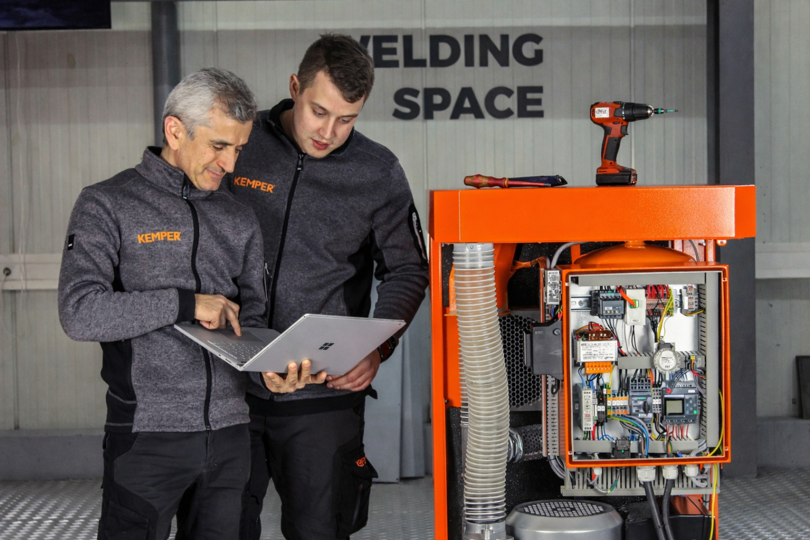 Zwei KEMPER Servicetechniker analysieren Anlagendaten auf einem Laptop neben einer geöffneten industriellen Absaug- und Steuerungseinheit.